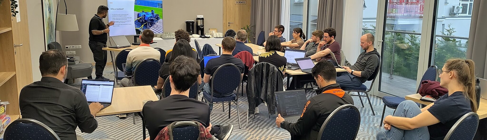 SQUARE research group at the Świnoujście retreat, 2026. We sit in a meeting room and listen to a presentation showing a BlueROV2 robot on the slide